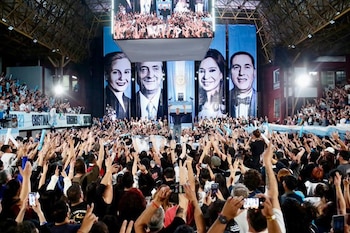 Un hombre en un escenario saluda a una multitud en un estadio cubierto decorado con cuatro retratos gigantes y una bandera argentina
