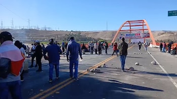 Puente de estructura naranja sobre una carretera con personas, rocas, humo y una pancarta en medio de un paisaje árido con postes de electricidad