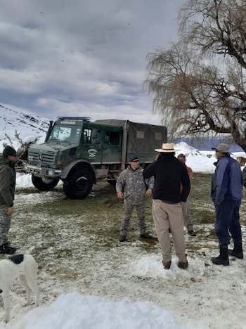 Camión verde de Gendarmería con hombres y un perro sobre terreno nevado y hierba. Montañas con nieve y árboles bajo cielo nublado en invierno