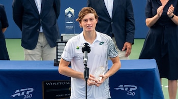 Un tenista joven de cabello castaño claro habla frente a un micrófono mientras sostiene un trofeo transparente en una pista de tenis.
