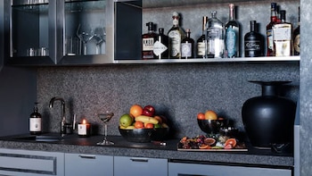 Encimera de cocina de piedra con grifo, botellas de licor en un estante, un cuenco con frutas, copas y una jarra negra