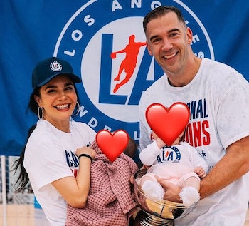 Martha Higareda y Lewis Howes sonríen a cámara, Higareda con un bebé envuelto en rosa y Howes sosteniendo otro bebé en un trofeo, con un logo deportivo de fondo