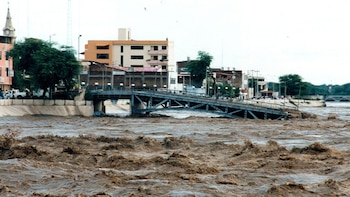 Río desbordado con agua turbulenta, un puente de metal colapsado, edificios de varios pisos, un campanario y personas en la orilla