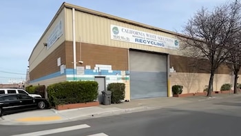 Fachada de un edificio industrial con un cartel de California Waste Solutions, una persiana metálica cerrada, plantas y vehículos estacionados