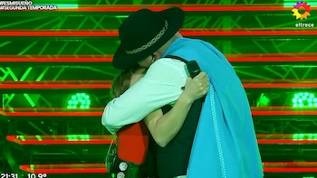 Un hombre con sombrero negro y poncho azul abraza a una mujer en un escenario iluminado con luces verdes