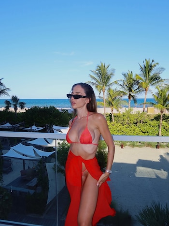 Una mujer en bikini rojo y pareo rojo posa en un balcón de vidrio. Al fondo se ve la playa, el mar y palmeras bajo un cielo azul despejado