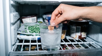Una mano coloca una moneda de euro sobre un bloque de hielo dentro de un vaso transparente situado en un congelador.