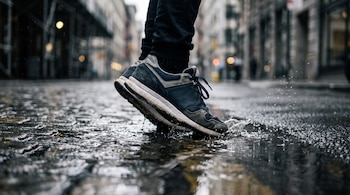 Primer plano de pies con zapatillas deportivas azules, grises y blancas pisando un charco en una calle mojada con edificios borrosos al fondo.