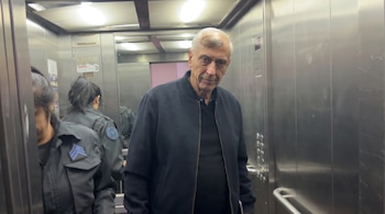 Un hombre de edad avanzada con chaqueta negra mira a la cámara dentro de un ascensor metálico, flanqueado por dos personas en uniforme de seguridad.