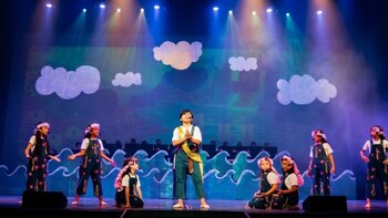 Un grupo de niños con overoles actúa en un escenario teatral decorado con figuras de nubes y olas marinas de utilería iluminadas