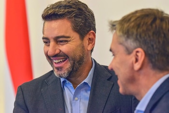 Primer plano de Leandro Zdero, un hombre de cabello oscuro y barba, sonriendo y mirando hacia la derecha, con un hombre parcialmente visible a su derecha y una bandera roja y blanca al fondo