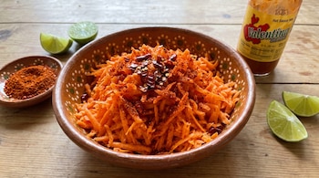 Un cuenco de cerámica con zanahoria rallada, chile en polvo y hojuelas de chile seco. Hay un plato pequeño con chile, limas y una botella de salsa picante.