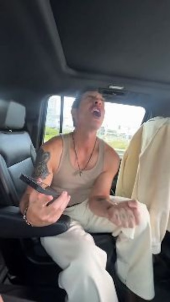 Hombre con camiseta sin mangas y pantalón blanco en el asiento trasero de un vehículo, sujetando un control remoto, con la boca abierta