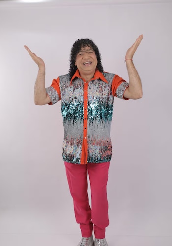 Hombre sonriente de cabello rizado oscuro, camisa de lentejuelas anaranjadas y plateadas, pantalones rosados y zapatillas, con los brazos levantados