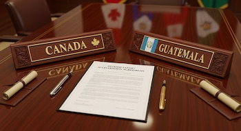 Mesa de madera con identificadores de Canadá y Guatemala, un documento de 'Defense Export Acceleration Agreement', dos plumas y dos pergaminos.