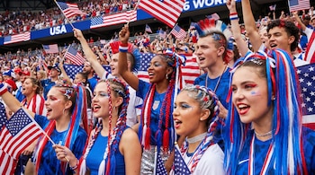 Un grupo de jóvenes con elaborados peinados rojos, blancos y azules ondean banderas de Estados Unidos en una tribuna de estadio llena de gente y banderas.