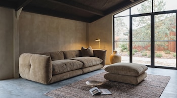 Un salón moderno con un sofá mullido de color beige, un otomán a juego, una alfombra texturizada y una lámpara de pie dorada. Un gran ventanal muestra árboles y un jardín.