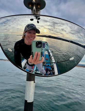 Reflejo de una persona sonriente con gorra tomando un selfi en el espejo de un mástil. Incluye el agua, un bote, remos, cielo nublado, sol y un anemómetro