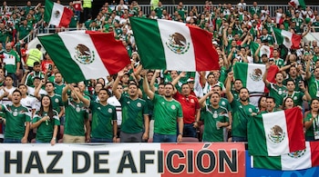 Una multitud de aficionados mexicanos, vestidos con camisetas verdes, ondean banderas de México en un estadio lleno. Se ve una pancarta con las palabras 'HAZ DE AFICIÓN'.