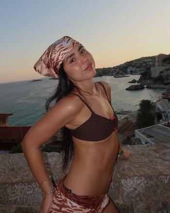 Una mujer joven con pañuelo, top de bikini y pareo posa sobre un muro de piedra, con una bahía, edificios y el mar al fondo