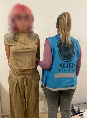 Una mujer de cabello rosado y vestimenta color caqui es custodiada por una agente de la Policía de la Ciudad, que viste un chaleco azul y se muestra de espaldas