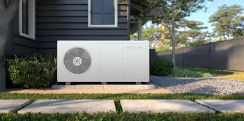 Una unidad blanca con ventilador y logo "XFRA" se ubica sobre una losa de hormigón al lado de una casa moderna de siding oscuro, con jardín y árboles al fondo