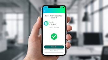 Una mano sostiene un teléfono móvil que muestra la pantalla de una aplicación bancaria con la confirmación de un envío de dinero exitoso.