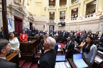 Vista interior de una sala grande con personas de pie en el centro y en balcones superiores, algunas con trajes formales. Hay una bandera argentina visible