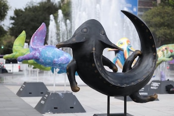 Escultura de bronce oscura con manchas circulares doradas. Al fondo, dos esculturas coloridas y una fuente de agua. Se ven árboles y cielo gris