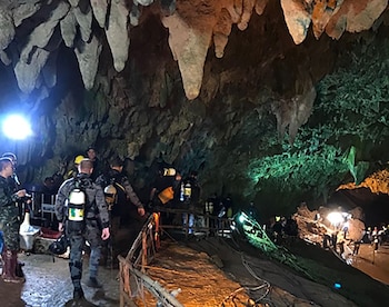 Interior de una cueva oscura con estalactitas, rocas, varias personas, algunas con trajes de buceo y tanques, luces, cables y pasamanos de madera