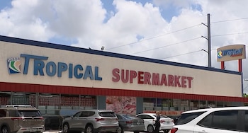 Vista exterior de la fachada del supermercado Tropical con su cartel azul y rojo, un estacionamiento con varios vehículos, y un cielo nublado