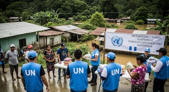Voluntarios de la ONU entregan ayuda a residentes en una comunidad guatemalteca con casas parcialmente inundadas. Un cartel de "Fondos de Emergencia ONU" es visible.