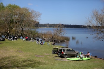 Personas sentadas en sillas de playa a la orilla de un lago junto a autos estacionados y un kayak verde en el pasto