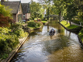 Un bote con dos personas navegando un canal en Giethoorn, flanqueado por casas de ladrillo y techos de paja, árboles verdes y un puente de madera al fondo