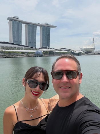 Hombre y mujer sonriendo con gafas de sol delante de un cuerpo de agua y un complejo de edificios modernos, incluyendo tres torres y una estructura blanca