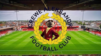 Estadio de fútbol con campo verde y gradas rojas. Un logo central muestra una suricata, corona de laurel y el nombre Crewe Aleksandr Football Club.