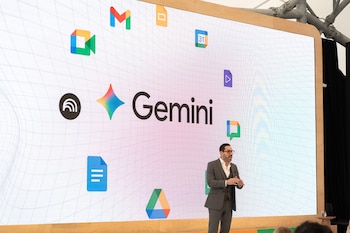 Hombre con traje oscuro y gafas presenta frente a una pantalla grande que muestra la palabra Gemini y varios iconos de aplicaciones coloridos sobre un fondo blanco cuadriculado