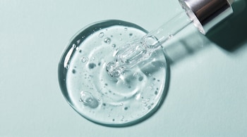 Vista cenital de una pipeta transparente que deposita una gota grande de gel transparente con burbujas sobre una superficie azul claro.