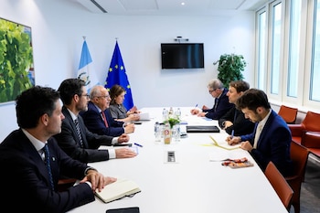 Vista lateral de una mesa de conferencias blanca donde ocho personas, vestidas formalmente, están sentadas. En el fondo, las banderas de Guatemala y la Unión Europea y una ventana