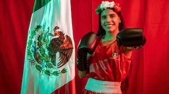 Una mujer con uniforme rojo de boxeo, guantes negros y corona de flores posa de frente junto a la bandera de México sobre un fondo rojo.