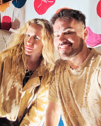 Julieta Prandi y un hombre con barba posan en primer plano con prendas de tono beige frente a una tela con formas de colores
