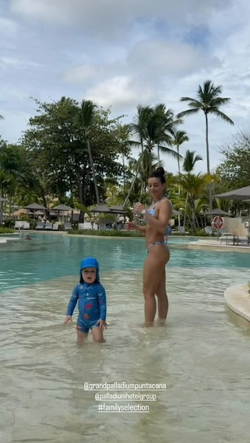 Sol Pérez en traje de baño junto a un niño con traje acuático azul dentro de una piscina rodeada de palmeras