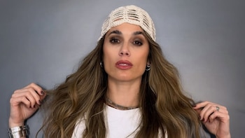 Retrato de Chechu Bonelli de frente, con cabello largo castaño claro, un gorro de punto color crema y maquillaje marcado. Toca su cabello con ambas manos
