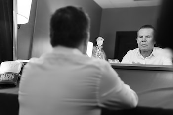 Julio César Chávez sentado de espaldas ante un espejo que muestra su rostro reflejado, junto a un guante de boxeo sobre la mesa