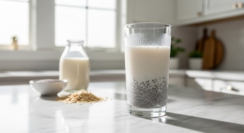 Vaso alto con agua de avena y semillas de chía sobre una barra de mármol blanco, con ingredientes como avena y leche en una cocina iluminada por el sol.