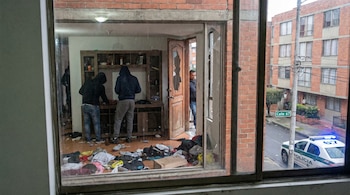 Vista a través de una ventana que muestra el interior de un apartamento desordenado con dos figuras encapuchadas, una puerta de madera rota y un vehículo policial en la Calle 67.