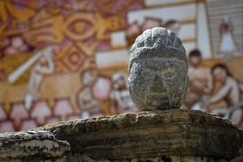 Primer plano de una escultura prehispánica de cabeza, tallada en piedra gris, apoyada sobre una estructura de piedra. Un mural ancestral desenfocado al fondo