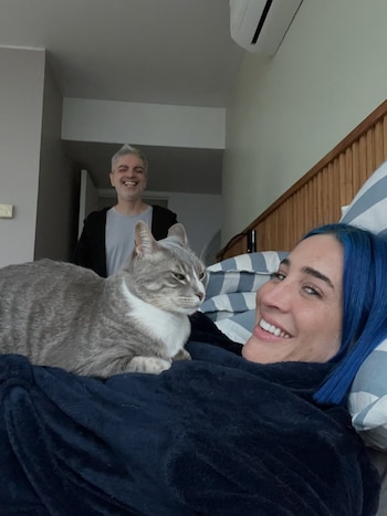 Una mujer de cabello azul está recostada en una cama con un gato gris en el pecho y un hombre con ropa gris y negra sonríe detrás