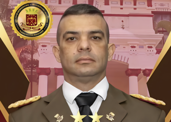 Retrato de busto de hombre con uniforme militar marrón, camisa blanca, corbata negra. Insignias doradas en solapas y hombros. Emblema dorado. Fondo arquitectónico