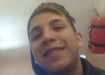 Primer plano de un joven sonriendo a cámara, con gorra oscura y ropa oscura. Se observa una pared clara y un objeto rojo difuso al fondo
